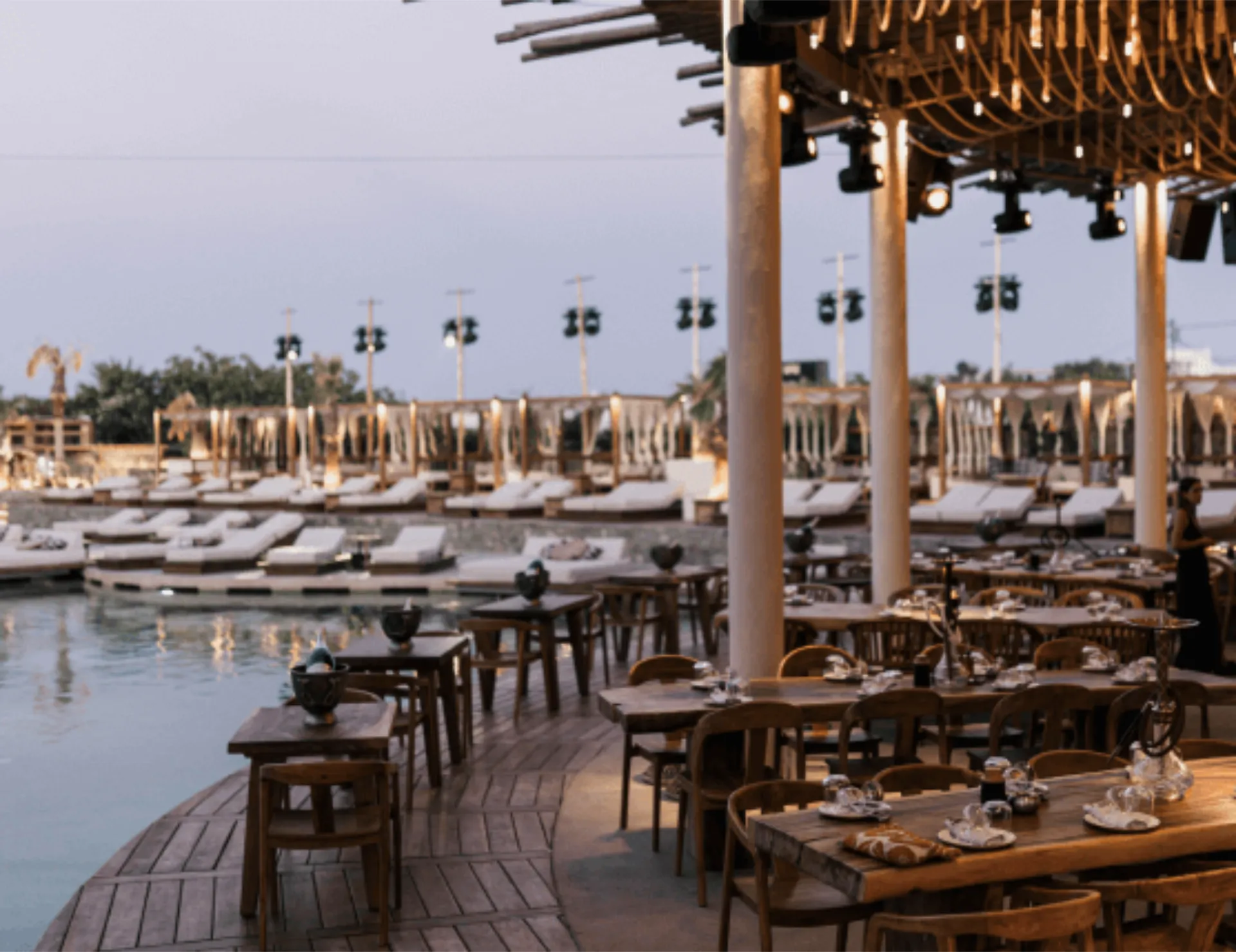 SantAnna's poolside restaurant with wooden tables set for dining overlooking lounge beds and cabanas at dusk.
