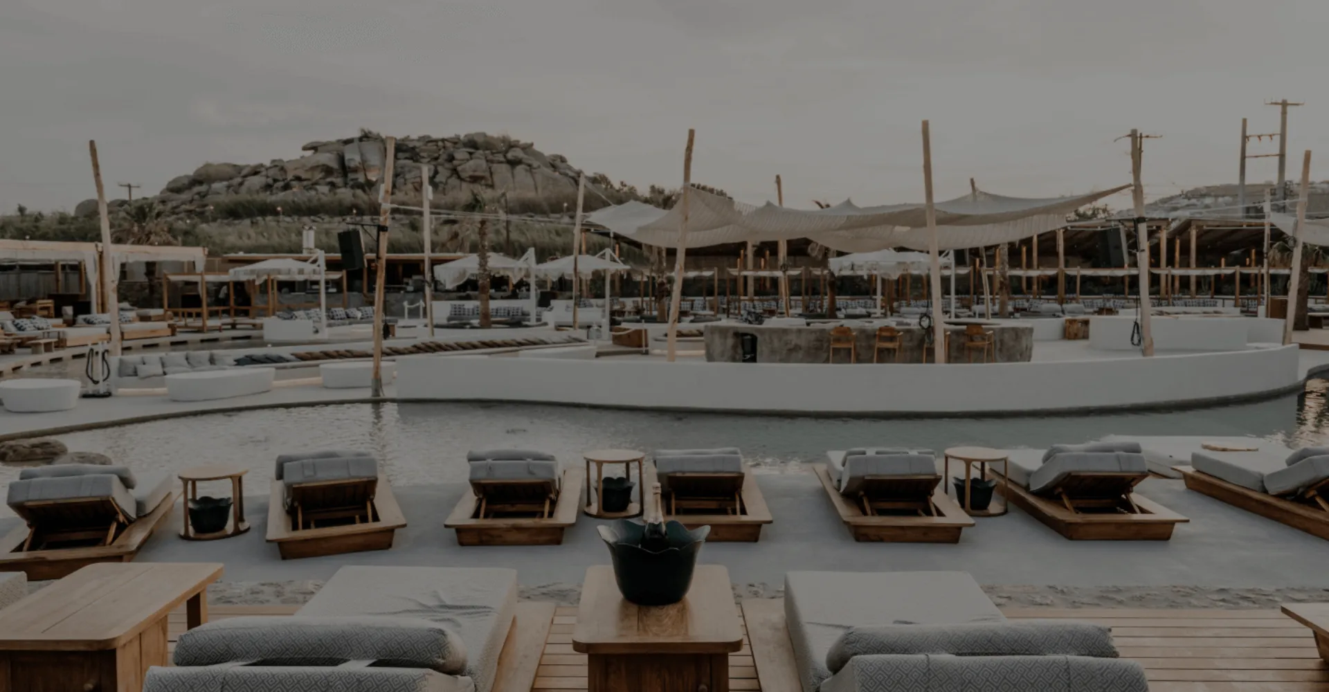 A panoramic view of SantAnna Mykonos Beach Club's main pool area at dusk. Wooden loungers with plush gray cushions line the shallow pool's edge, each with small side tables. The expansive space features minimalist white architecture with natural wooden posts supporting canvas canopies. In the background, rocky Mykonian hills rise against a muted gray sky, creating the signature Cycladic landscape. The serene setting combines luxury beach club amenities with the natural beauty of the island's terrain.